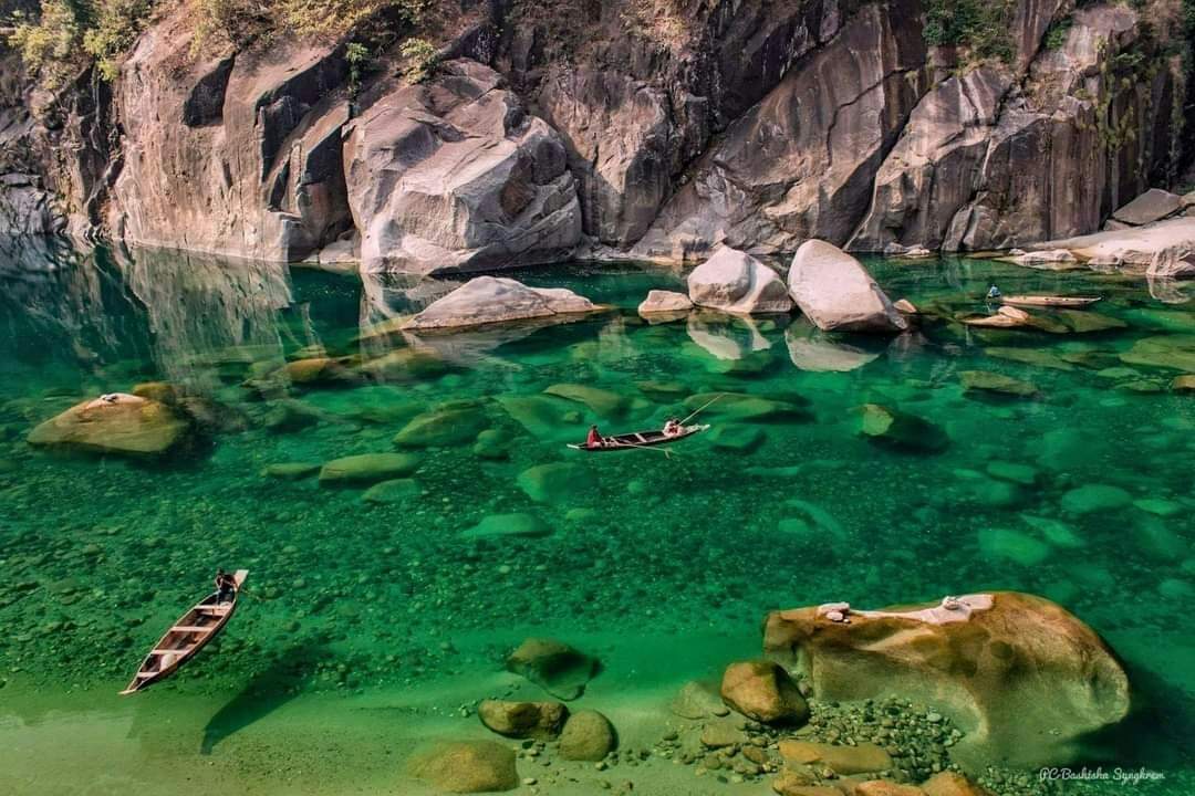 Crystal-clear emerald waters of the Umngot River in Dawki, Meghalaya, with wooden boats floating above the visible riverbed stones.
